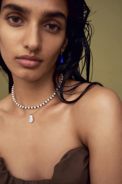 Close-up of a woman with dark hair wearing pearl jewelry and a brown strapless top against a neutral backdrop in a studio.