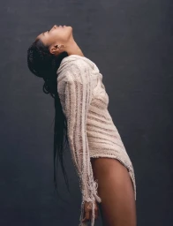 Portrait of a woman with long dark braids leaning backward wearing a textured cream bodysuit against a dark grey textured background.