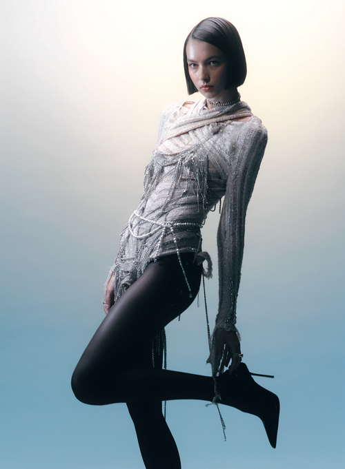 A fashionable young woman poses indoors, wearing a white top, a beaded shawl, black tights, and high heels in a studio setting.