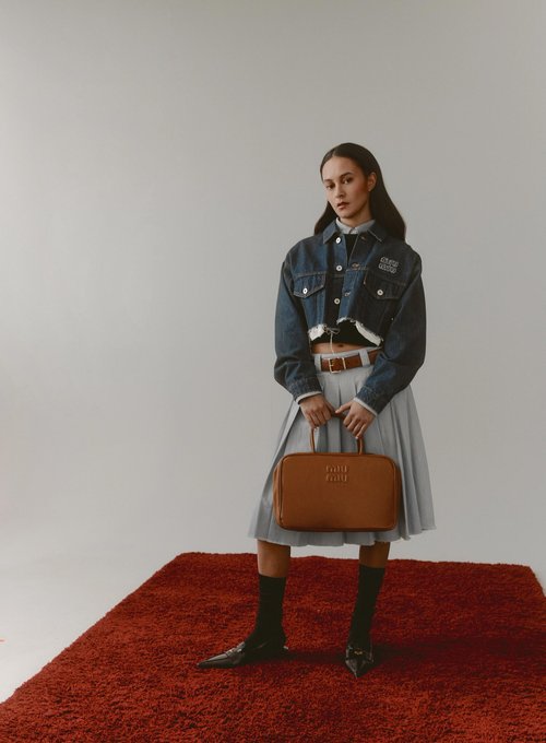 A young woman in a cropped denim jacket and grey pleated skirt poses with a brown satchel purse on a red shag rug.