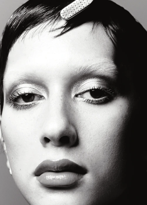 Close-up portrait of a woman with short dark hair, bleached brows, and glossy lips against a neutral background.