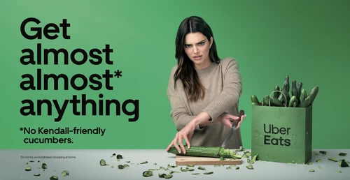 Uber Eats advertisement features Kendall Jenner awkwardly chopping a cucumber with many cucumbers in a bag and food delivery service logo.