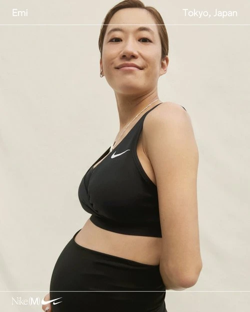 Portrait of a pregnant woman in black activewear, smiling indoors against a neutral background. Nike sports bra and leggings.