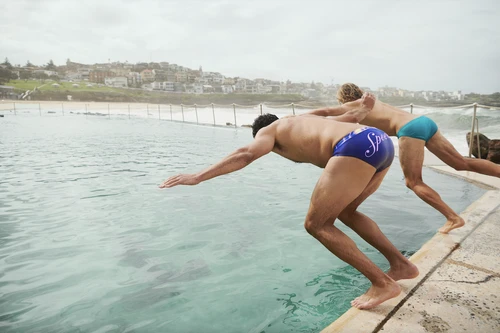 Two male swimmers dive into an ocean pool with a coastal cityscape visible in the background, evoking sport and outdoor recreation.