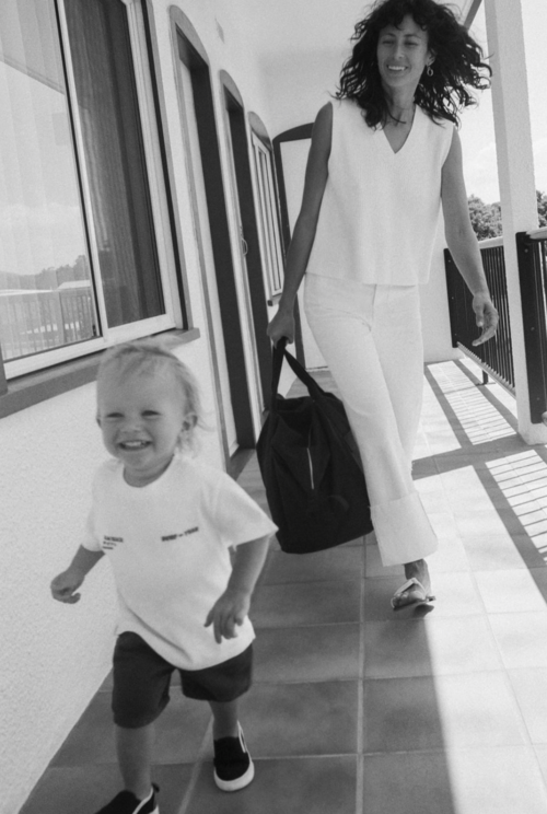 A mother and toddler walk down a corridor. The child smiles at the camera as the mother carries a tote bag in black and white.