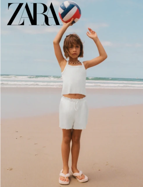A young girl on a beach with a volleyball, wearing a white tank top and shorts, standing near the ocean on a sunny day.
