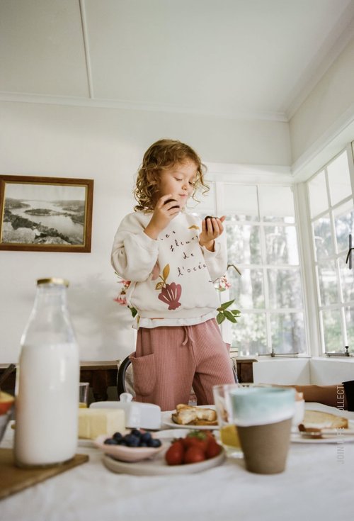 Young girl with curly hair eating a plum at a table with various breakfast foods, including strawberries, blueberries, and milk indoors.