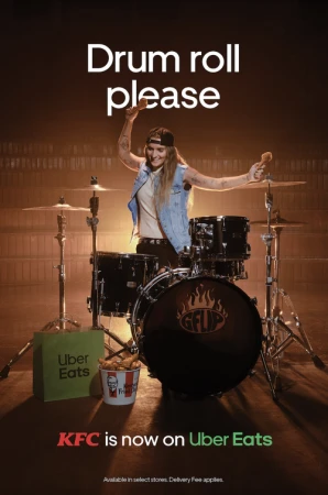 A young female drummer holds drumsticks in front of a drum kit next to a KFC bucket and Uber Eats bag in a promotional ad.
