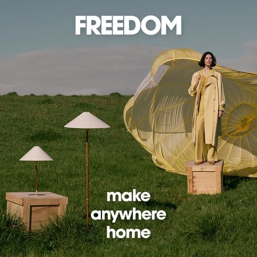 Woman in a yellow suit stands near a yellow parachute on a grassy field next to two standing lamps, evoking a surreal and whimsical scene.