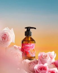 Amber glass bottle of body oil surrounded by light pink roses against a sunset gradient background