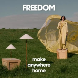 Woman in yellow trench coat standing on a wooden crate in a grassy field with lamps and a parachute structure