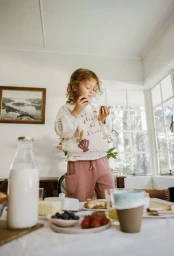A young child standing at a breakfast table filled with fruit, toast, and milk, holding blueberries