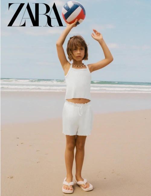 Young child holding a volleyball on a sandy beach wearing a white top and shorts, produced by Matt Chee.