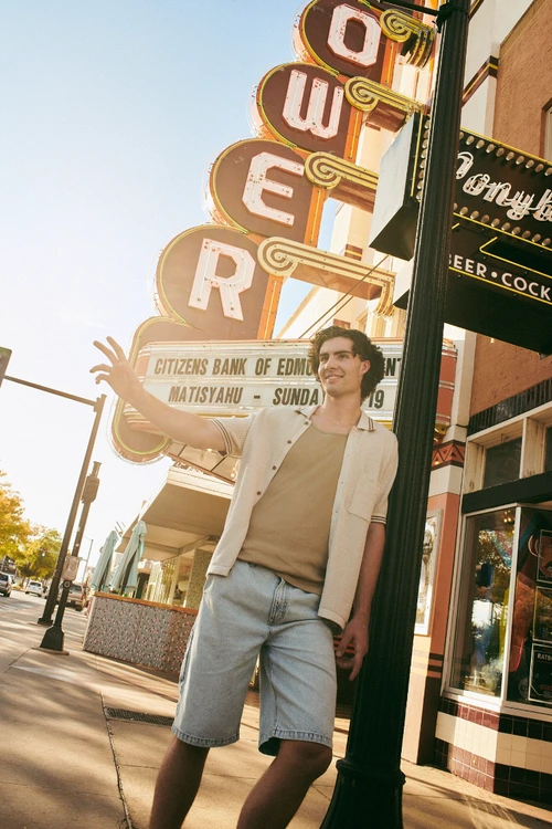 Young man in short-sleeve shirt and denim shorts leaning against a streetlamp beneath a vintage theater sign in Sydney