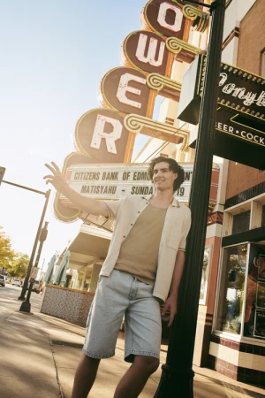 Young man in short-sleeve shirt and denim shorts leaning against a streetlamp beneath a vintage theater sign in Sydney