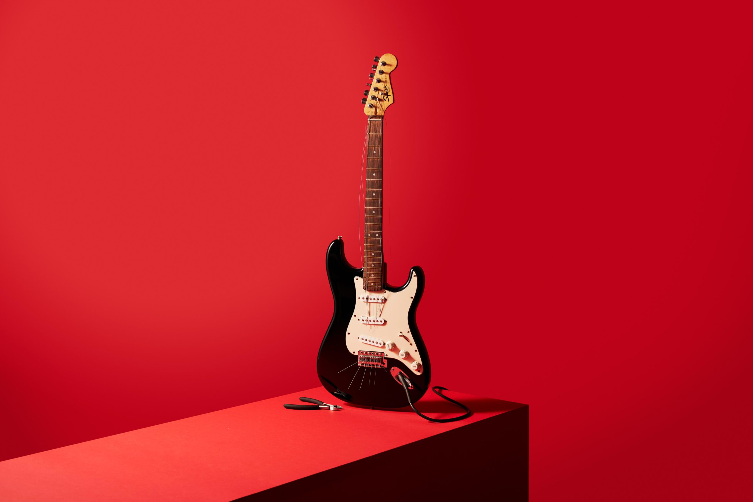 Electric guitar on a red cube against a red backdrop. The guitar is black and white, with a cable trailing on the cube.