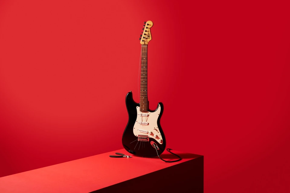 Electric guitar on a red cube against a red backdrop. The guitar is black and white, with a cable trailing on the cube.