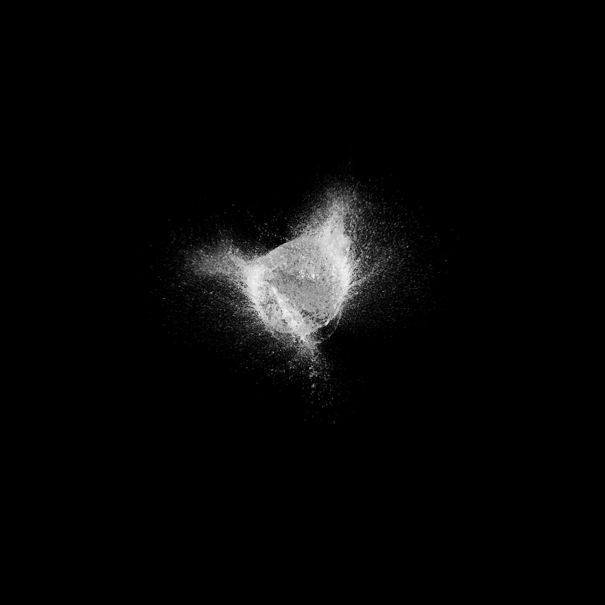 Dynamic monochrome image of a burst of white powder suspended in mid-air against a stark black backdrop, resembling an explosion or splash.