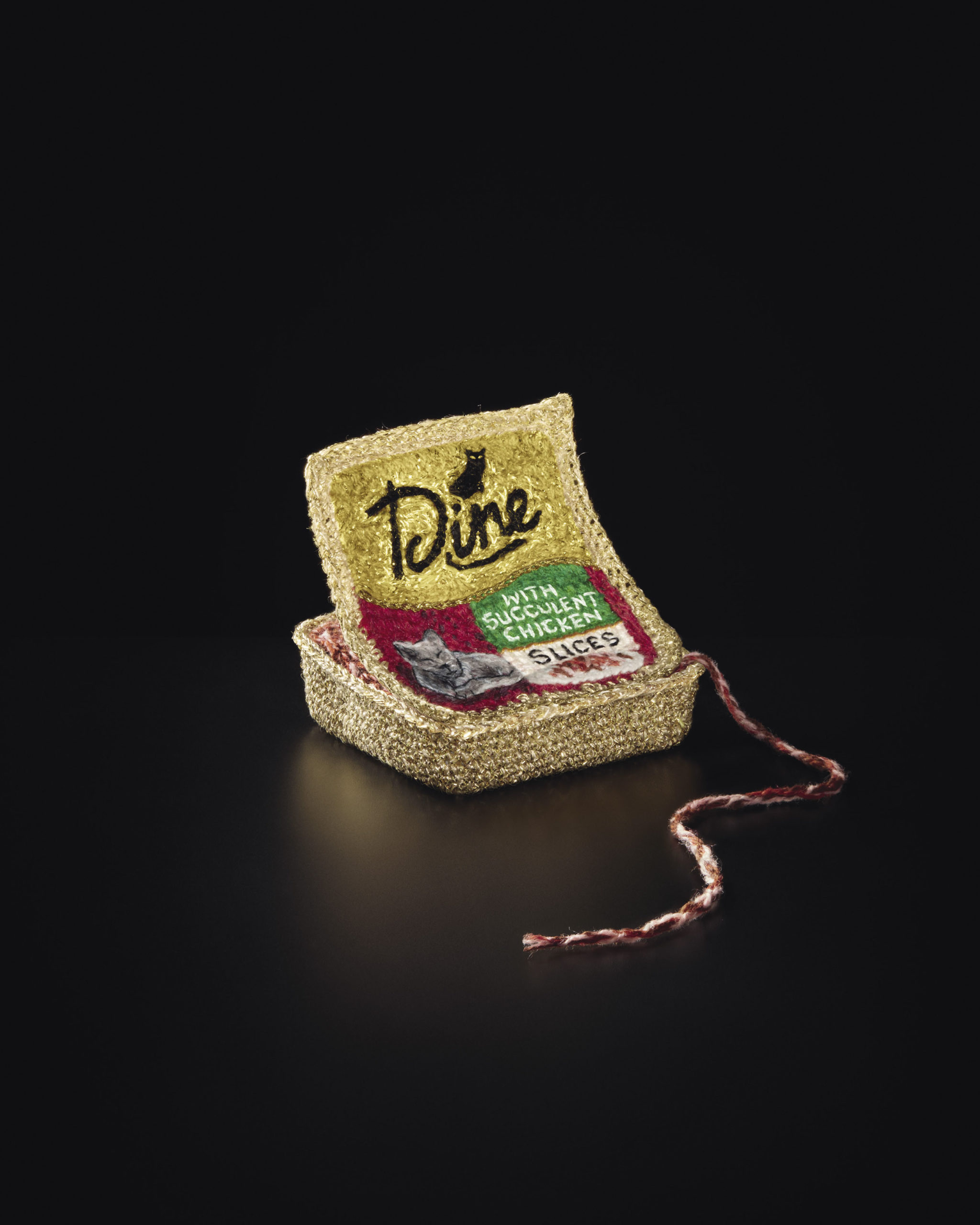Close-up of crocheted cat food can with gold, red, green and gray yarn details isolated on a black background with string detail.