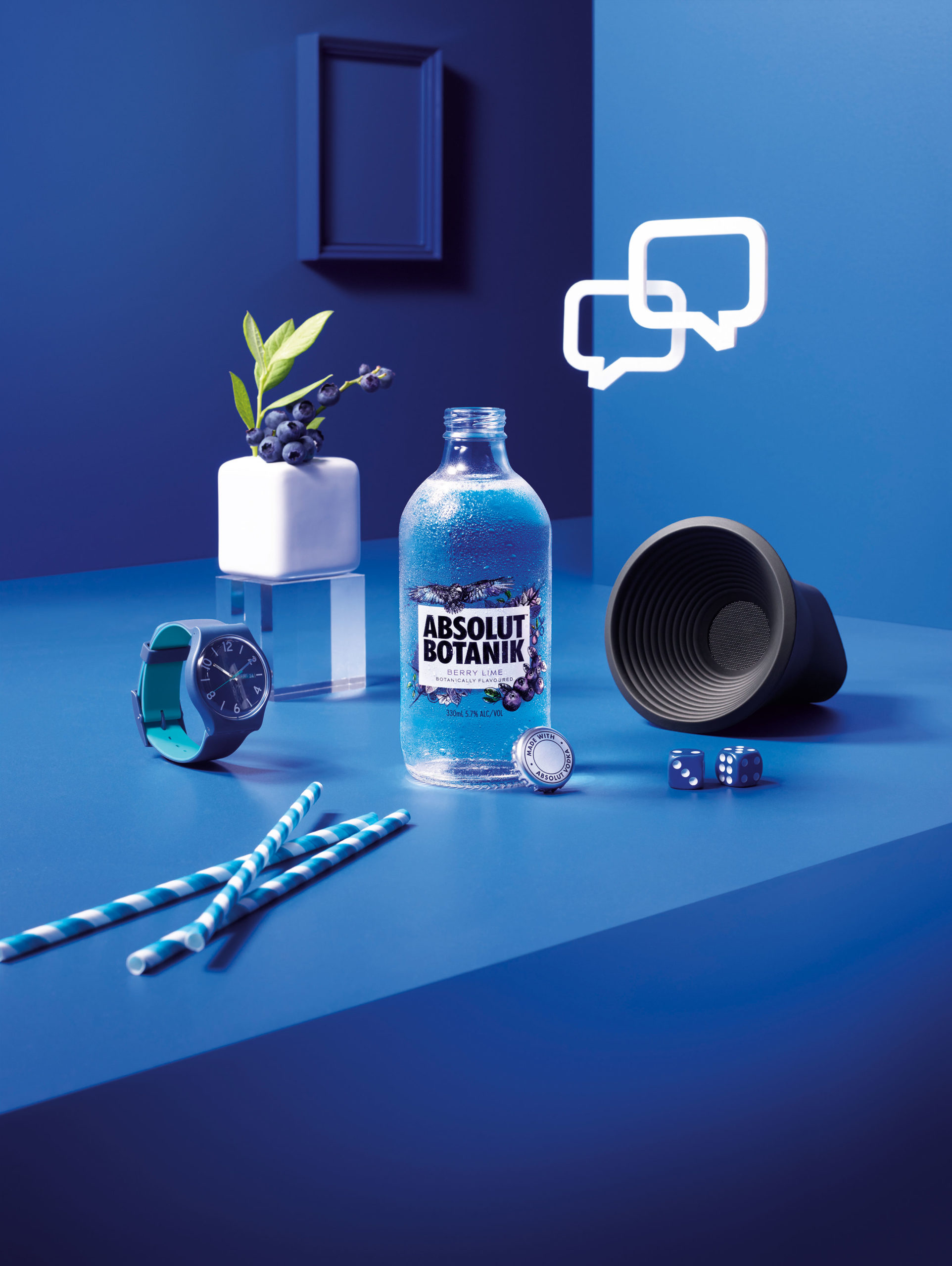 Still life of Absolut Botanik drink bottle with dice, speaker, watch, straws and blueberries against a vibrant blue background.