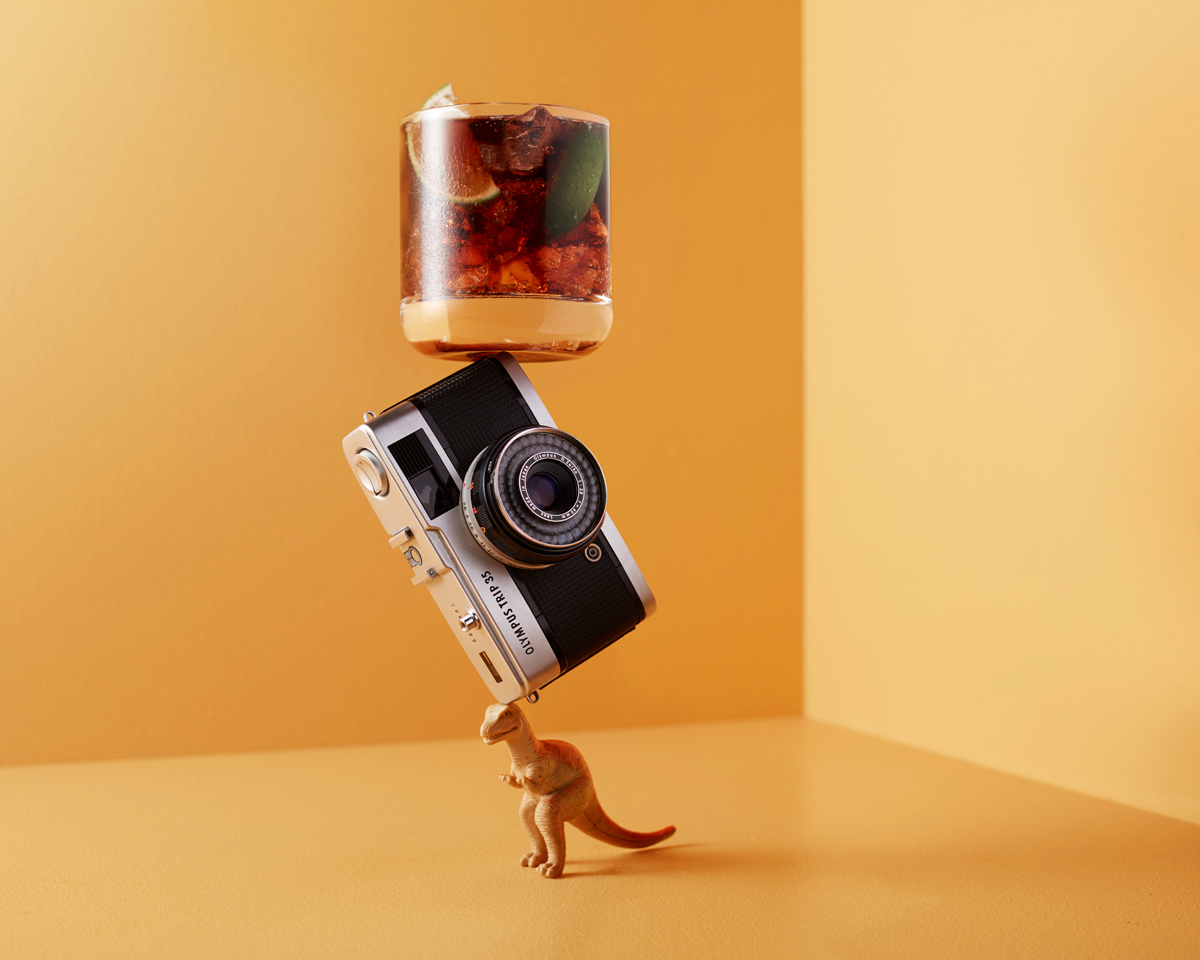 Retro camera balanced between a glass of mixed drink and a dinosaur toy against a neutral yellow backdrop in a studio setting.