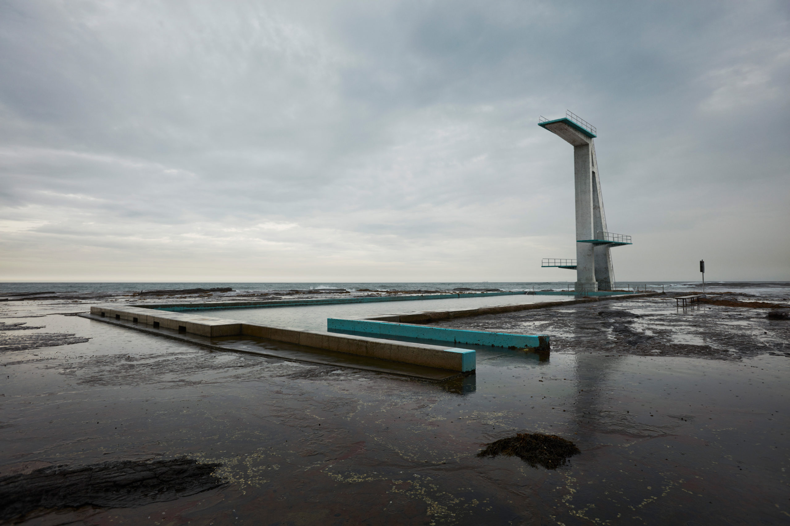 Outdoor swimming pool complex featuring diving platforms by the sea, under a cloudy sky in a landscape orientation. Water and rock.