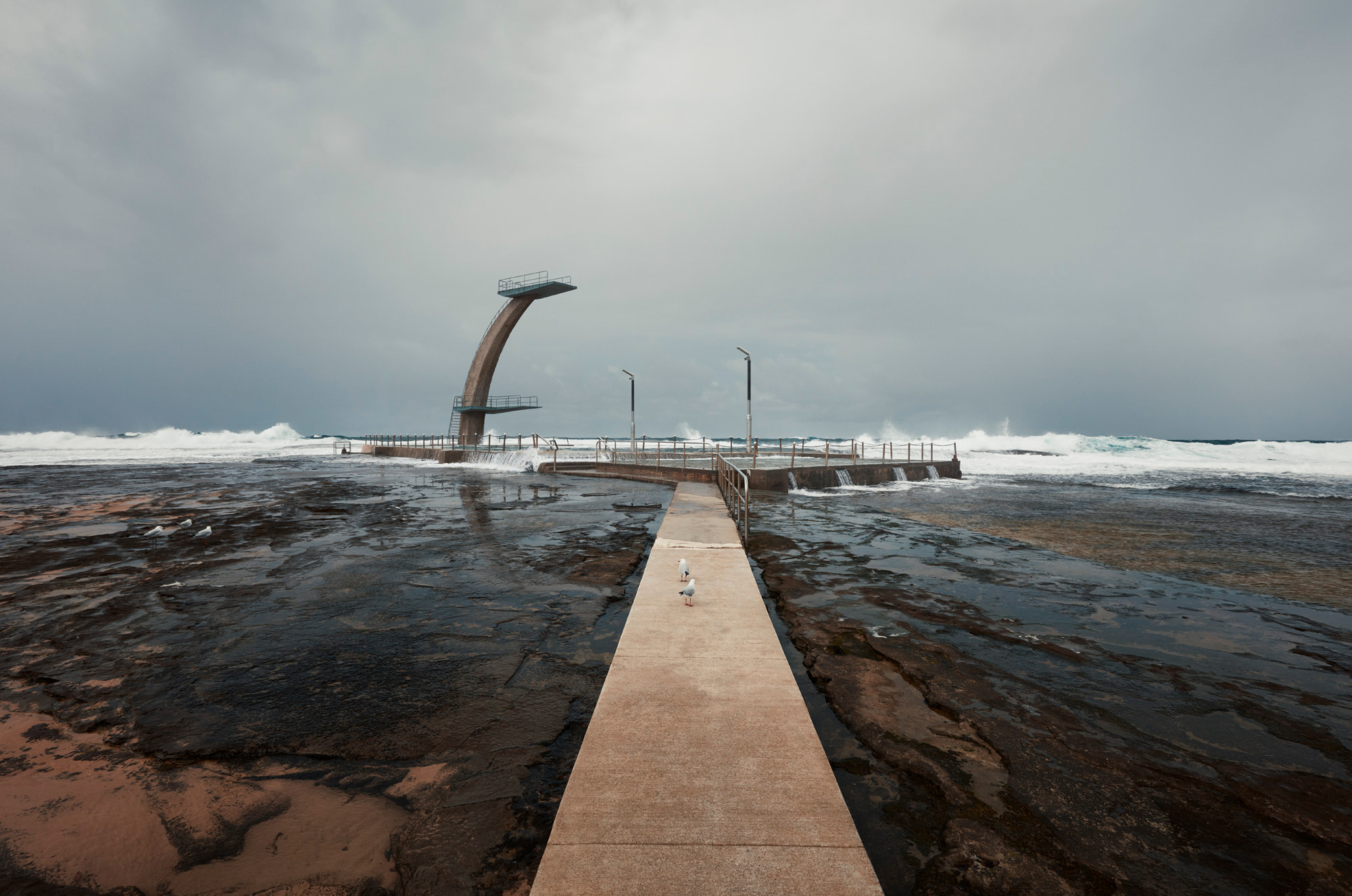 Ocean pool with diving platform and breaking waves against a cloudy sky, featuring a concrete walkway and several seabirds.