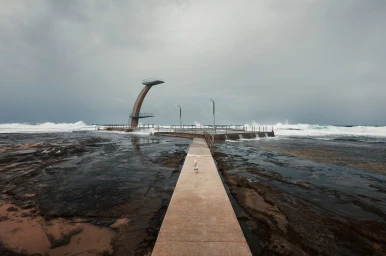Concrete walkway leading to an ocean pool with a curved diving tower under an overcast sky, captured in Sydney.