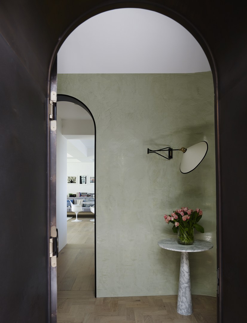 Interior view through an arched doorway with a round marble table, flowers, and wall lamp against a muted green wall.