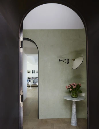 Interior view through an arched doorway with a round marble table, flowers, and wall lamp against a muted green wall.
