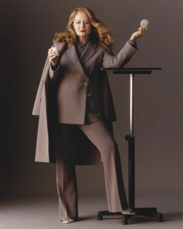 Elegant woman in a stylish suit holding a compact and drink in a studio setting, leaning on a black adjustable table.