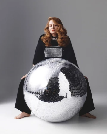 Woman with long auburn hair wearing black clothing seated behind a large silver disco ball against a grey background.