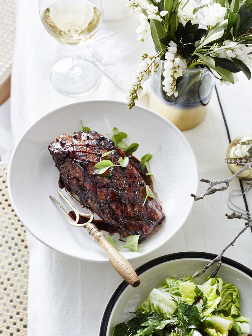Close-up of glazed meat dish with golden fork, white wine, and salad on a white tablecloth, illuminated by bright light.