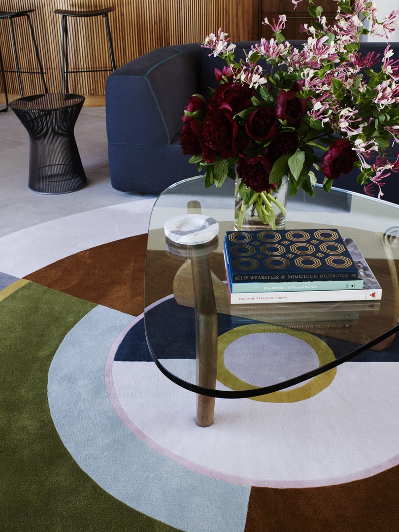 Still life shot of a glass coffee table with a flower arrangement and books, set on a colorful geometric rug with a sofa in the background.