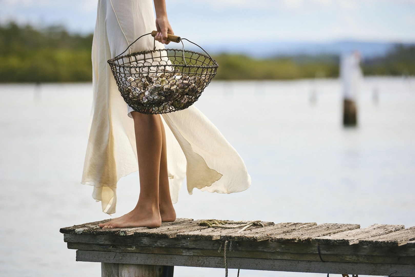 Woman in a white dress stands on a wooden dock holding a wire basket filled with oysters in a serene waterside setting.