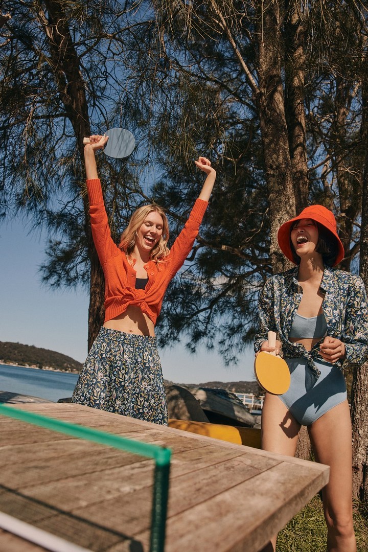Two women play paddle tennis on a wooden table outdoors, wearing casual outfits on a sunny day near a body of water.