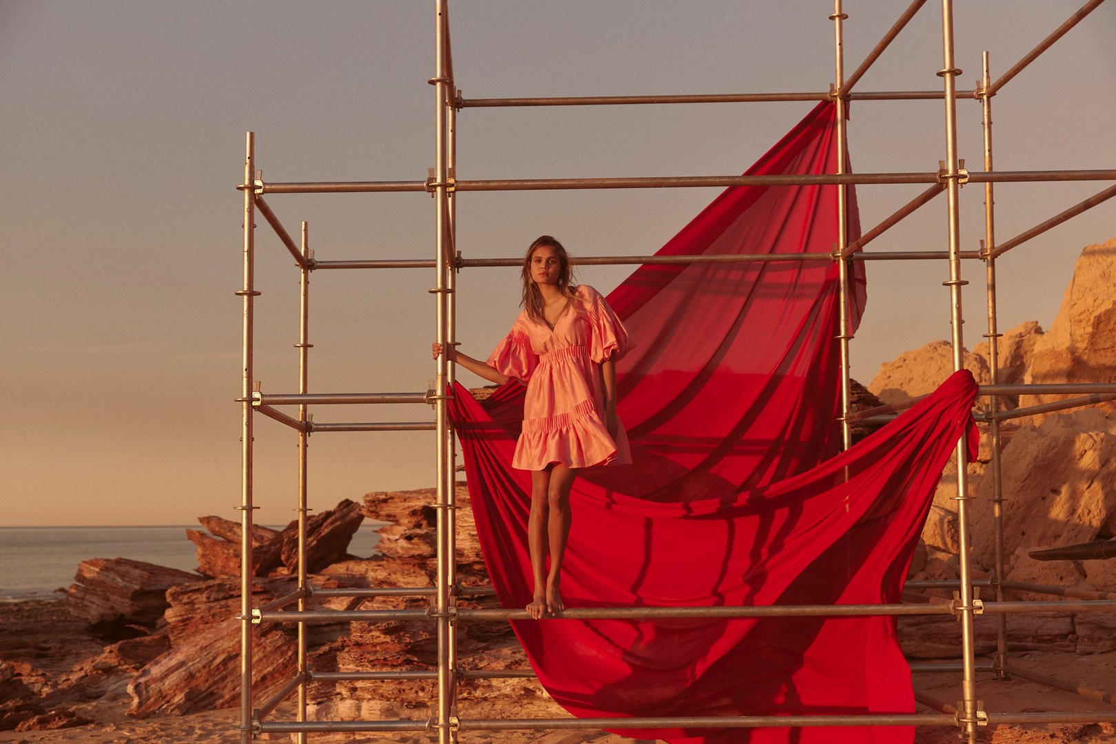 A barefoot woman in a pink dress stands on metal scaffolding by a red sheet on a rocky shore under an orange sky near the ocean.