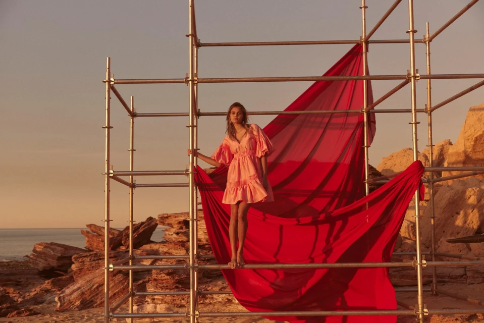 A barefoot woman in a pink dress stands on metal scaffolding by a red sheet on a rocky shore under an orange sky near the ocean.