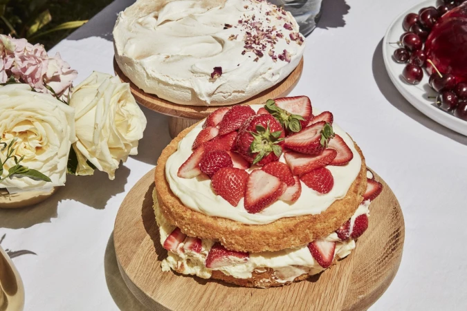 Overhead shot of a layered strawberry cake, meringue cake, cherries, and white roses displayed on a white tablecloth outdoors.