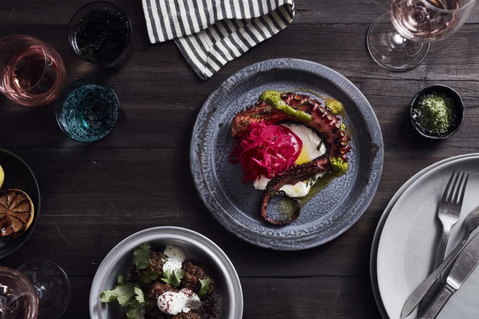 Overhead shot of octopus with white sauce and red onion on gray plate with glasses, silverware, and greens on a dark wooden table.