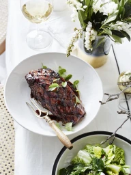 Glazed roasted meat dish served in a white bowl with a golden serving fork, accompanied by a glass of wine and salad.