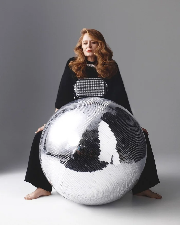 Woman with long auburn hair wearing black clothing seated behind a large silver disco ball against a grey background.