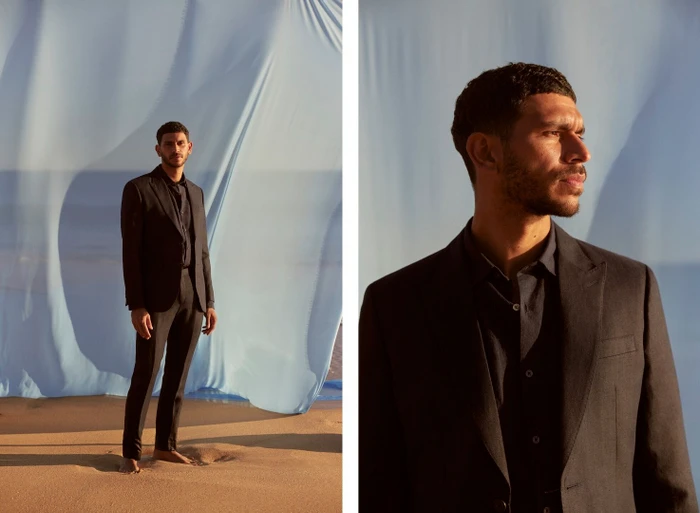 Man in a dark suit standing barefoot on sand before a large light blue fabric backdrop styled by Kirsten Bookallil