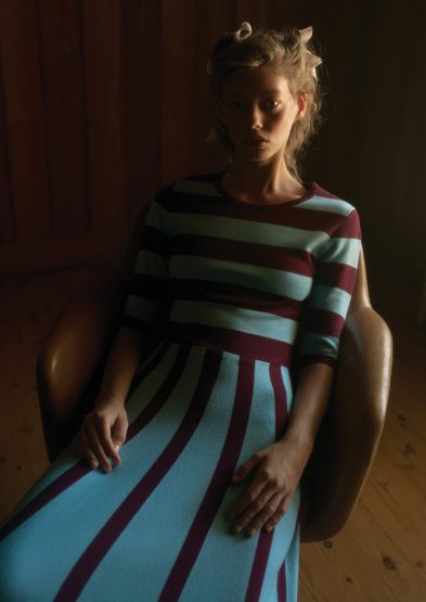 Woman with blonde hair in curlers sits in a chair wearing a striped turquoise and maroon dress in a dimly lit room with wood walls.
