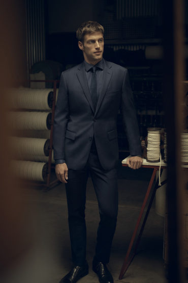 Full-length shot of a light-skinned adult man in a dark suit and tie, posing indoors with a serious expression in dim lighting.