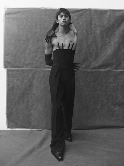 Monochrome studio shot of a young adult in a corset and dress pants with gloved arms reaching around them in a portrait.