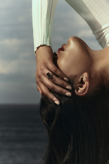 Close-up portrait of a woman lying down with her hand near her face, showcasing an elegant ring and a serene expression with ocean view.
