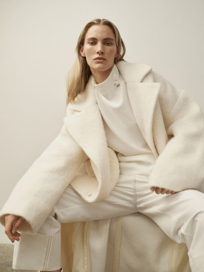 A blonde woman in a white coat and pants sits indoors against an off-white background, posing in fashion photography with neutral expression.