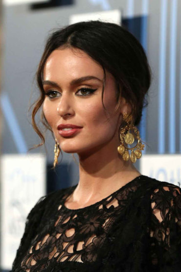 Elegant brunette woman in lace dress and gold earrings poses indoors at an event with a blurred background.