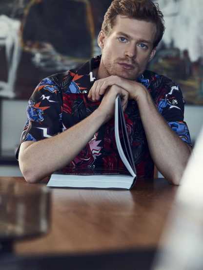 Red-haired man in floral shirt leans on book at wooden table with blurred background in eye-level portrait orientation indoors.